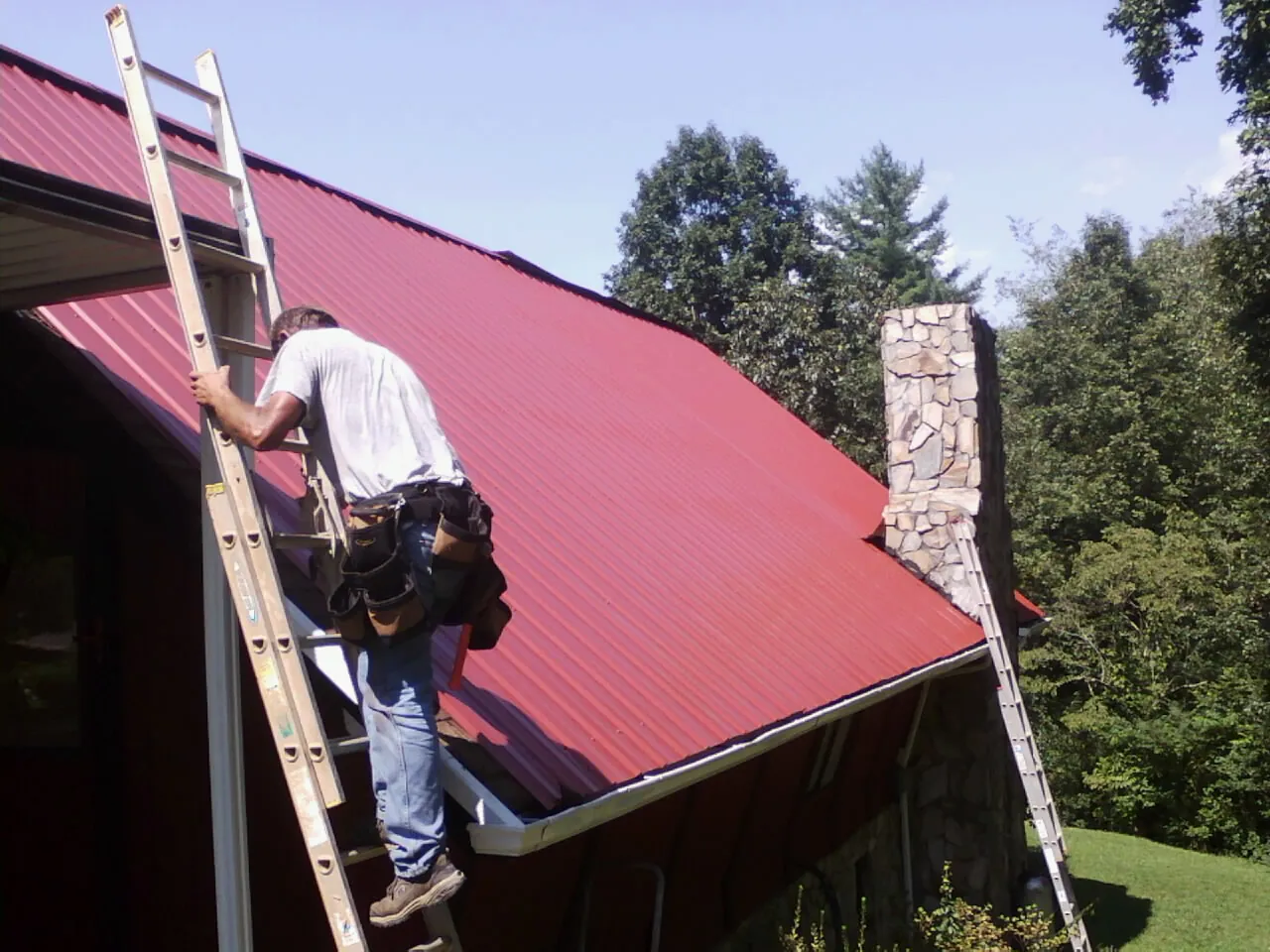 Roofer on a ladder beside a red metal roof and stone chimney — Soffit & Fascia Repair by Roofing Brothers in Bartlett